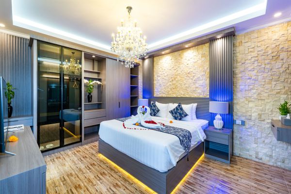 Luxurious hotel bedroom with king-sized bed scattered in rose petals, crystal chandelier, stone accent wall, glass wardrobe, and ambient blue LED lighting on wooden floors.